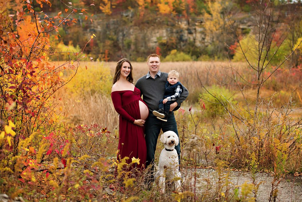 A mother to be iin a red maternity gown stands with her husband holding their toddler son and their white puppy in a park path
