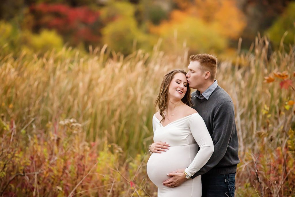 A mother to be stands in a field of tall grass and fall colors in a white maternity gown while being kissed and hugged by her partner in a grey sweater toronto doulas