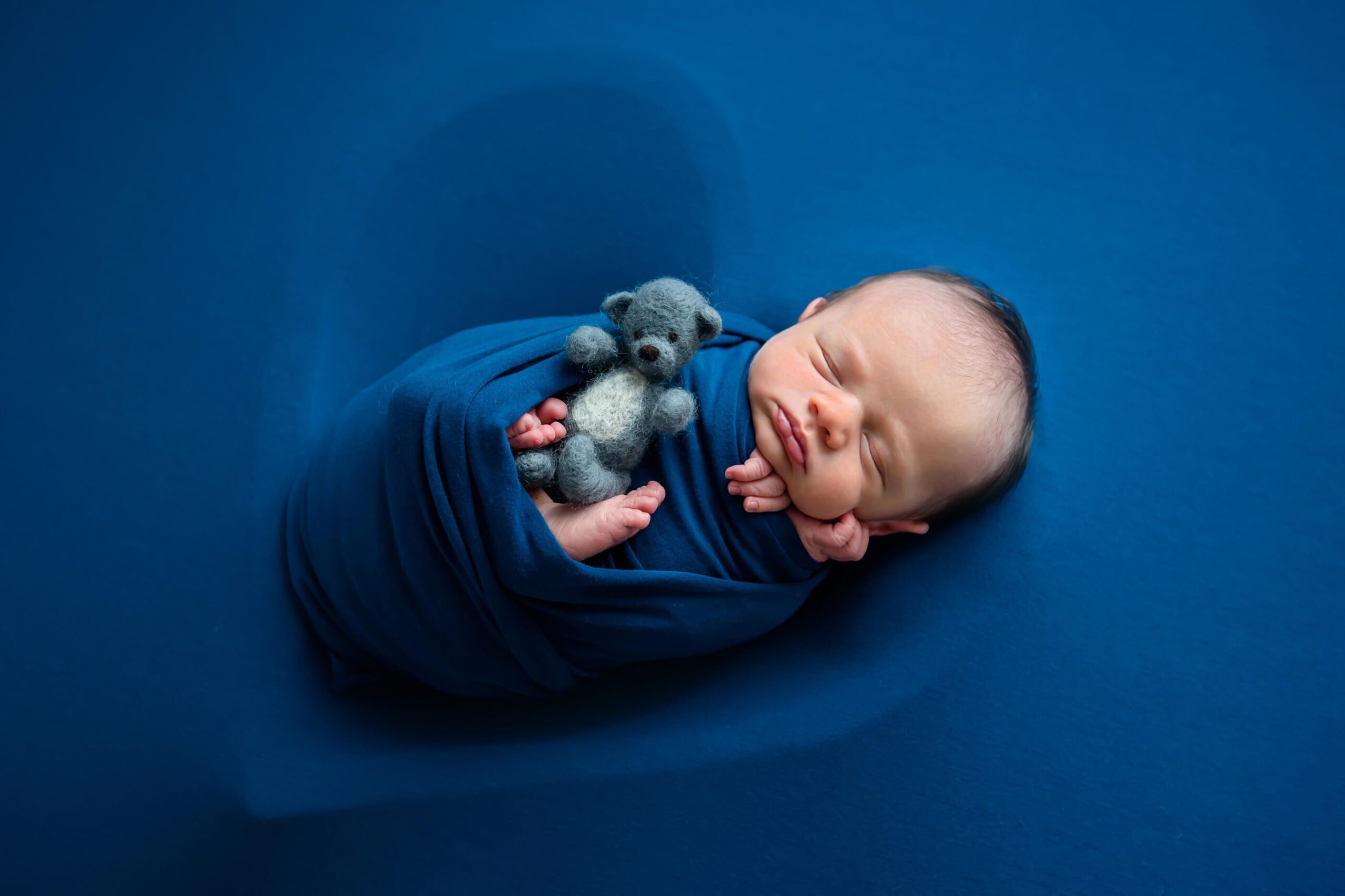 Newborn baby boy in a blue wrap with a heart bowl and a little teddybear.