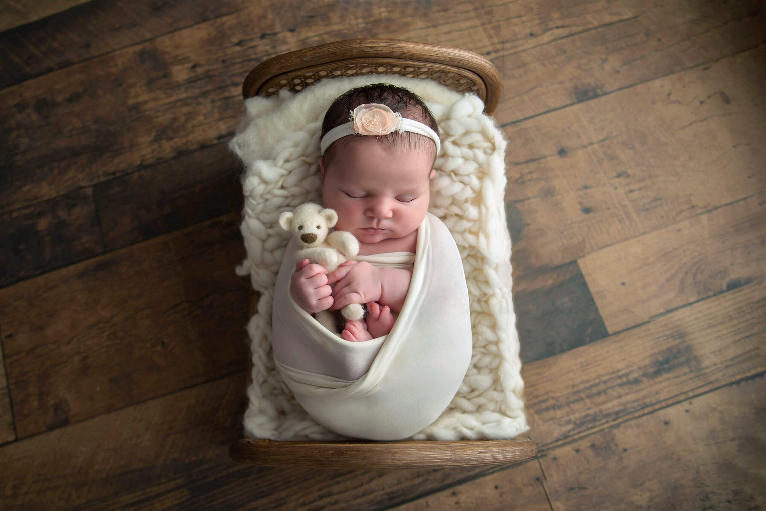 newborn girl in white wrap and white headband holding a teddy bear in a prop bed