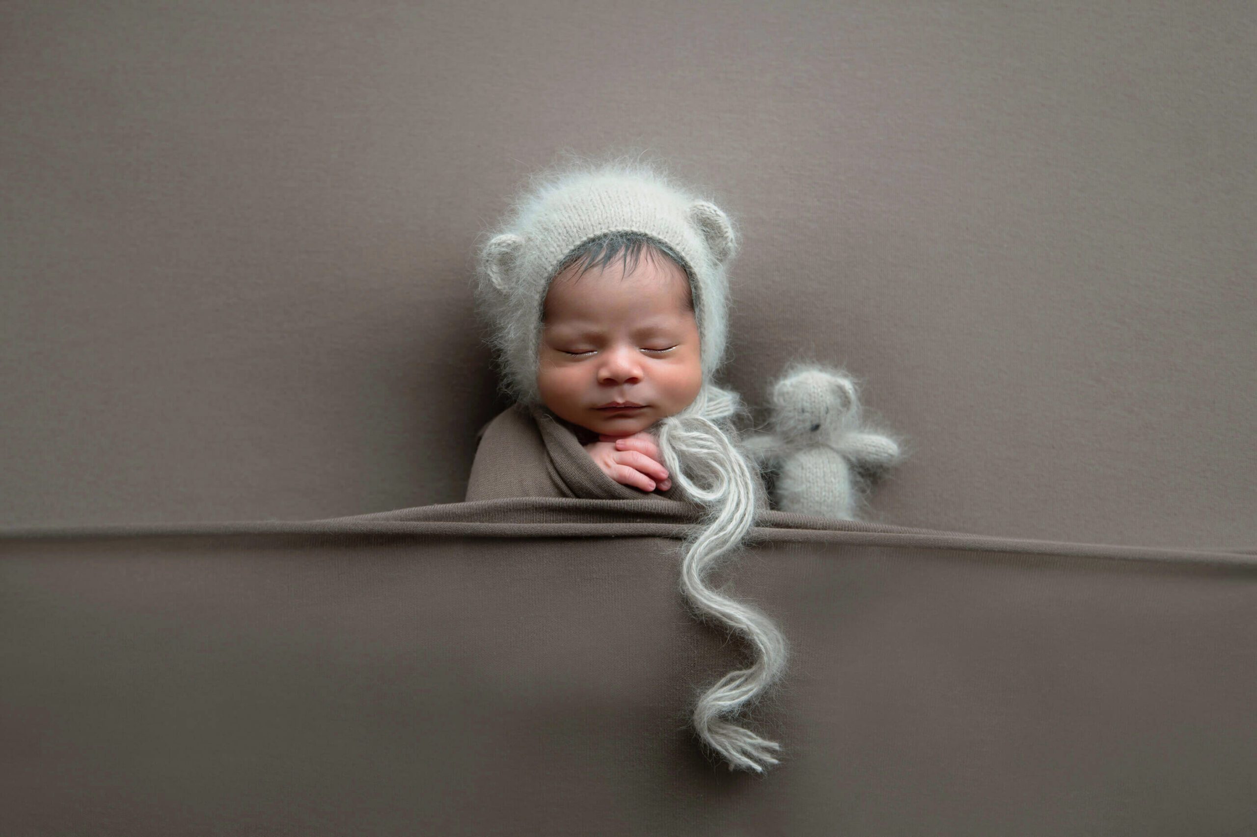 Newborn boy with bear hat and tiny bear laying down on the brown backdrop