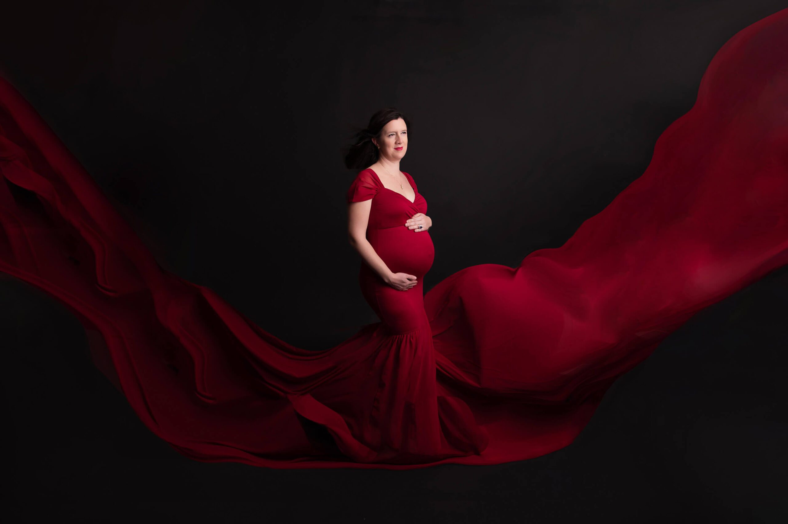 Black background studio maternity session mom in red dress flowing cradling belly