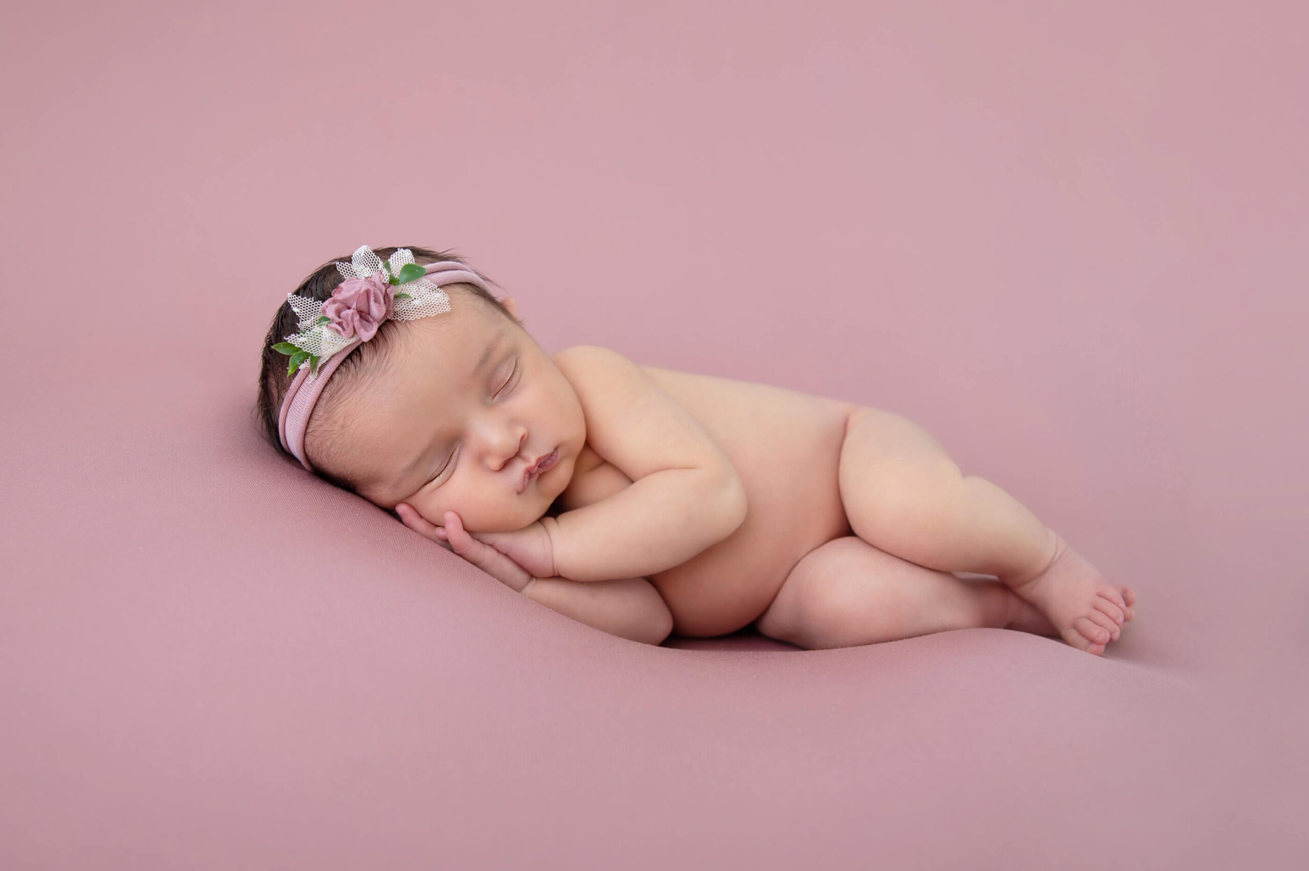 Newborn baby girl laying on her side with a pink headband on a pink background. Burlington Newborn Photographer