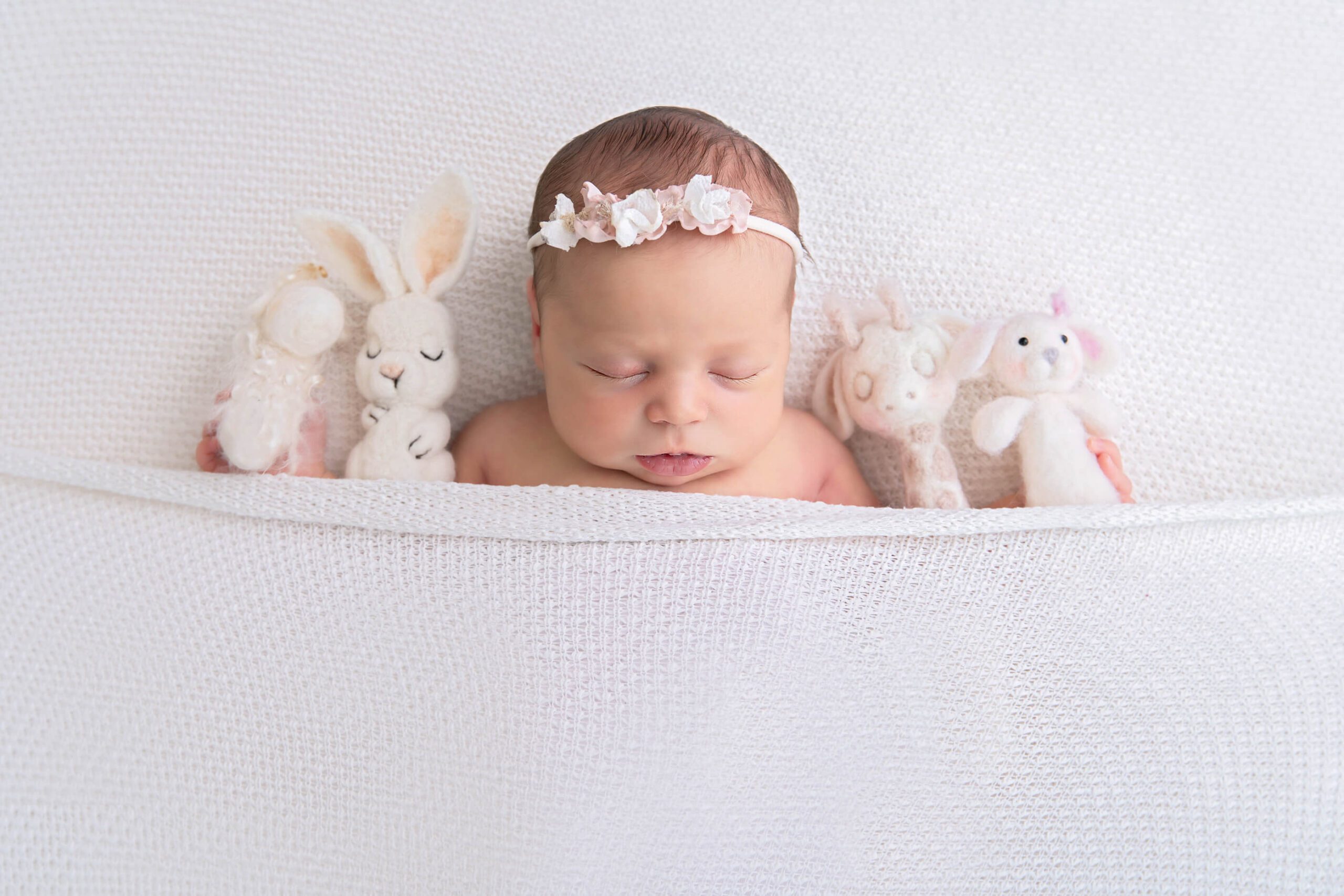 Newborn girl laying down in all white with a headband surrounded by stuffies.  Toronto Birth Centre