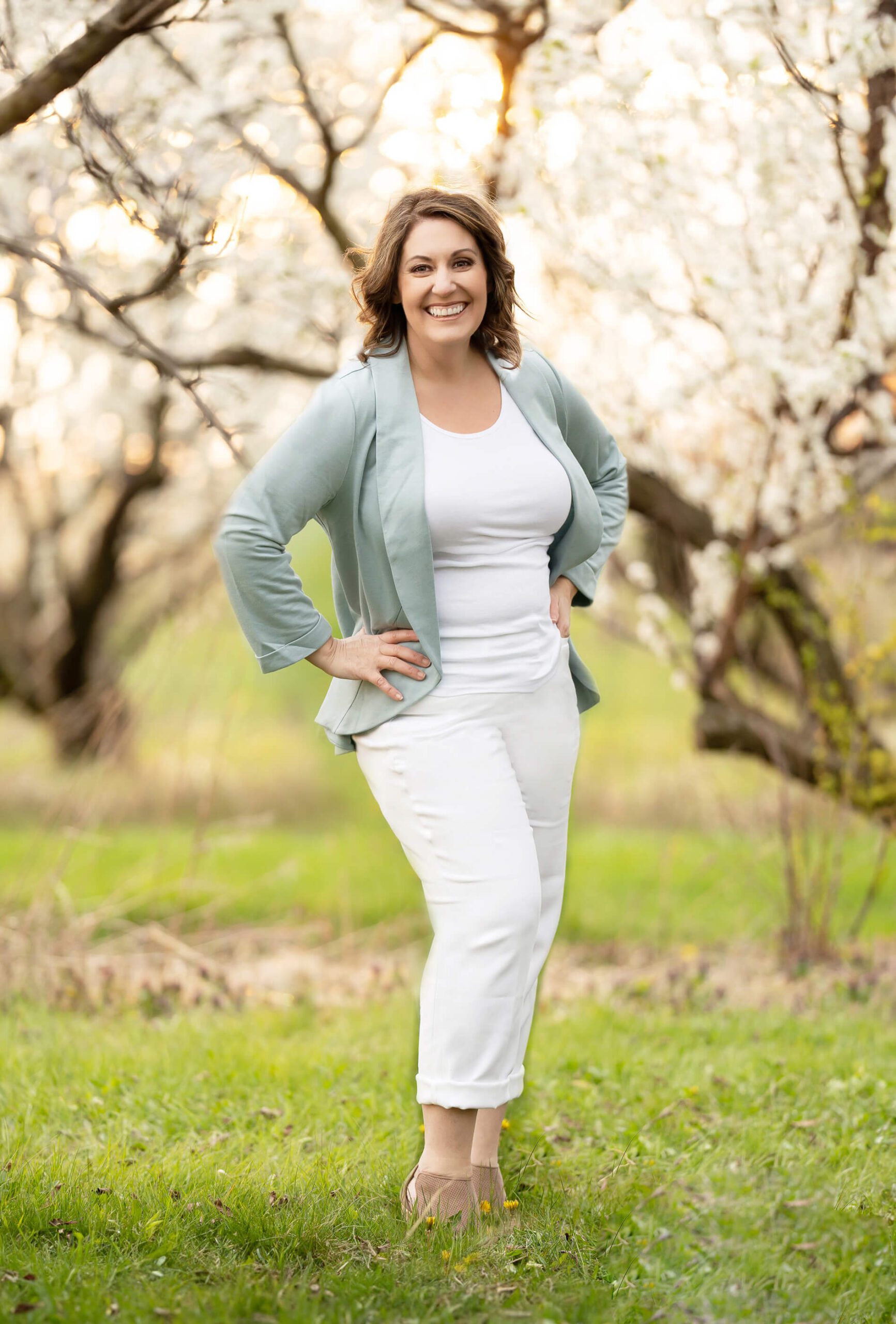 Photo of Amanda Gibson of Gibson Photography Toronto Newborn Photographer in the Cherry Blossoms wearing white pants and a blue top smiling with her hands on her hips.