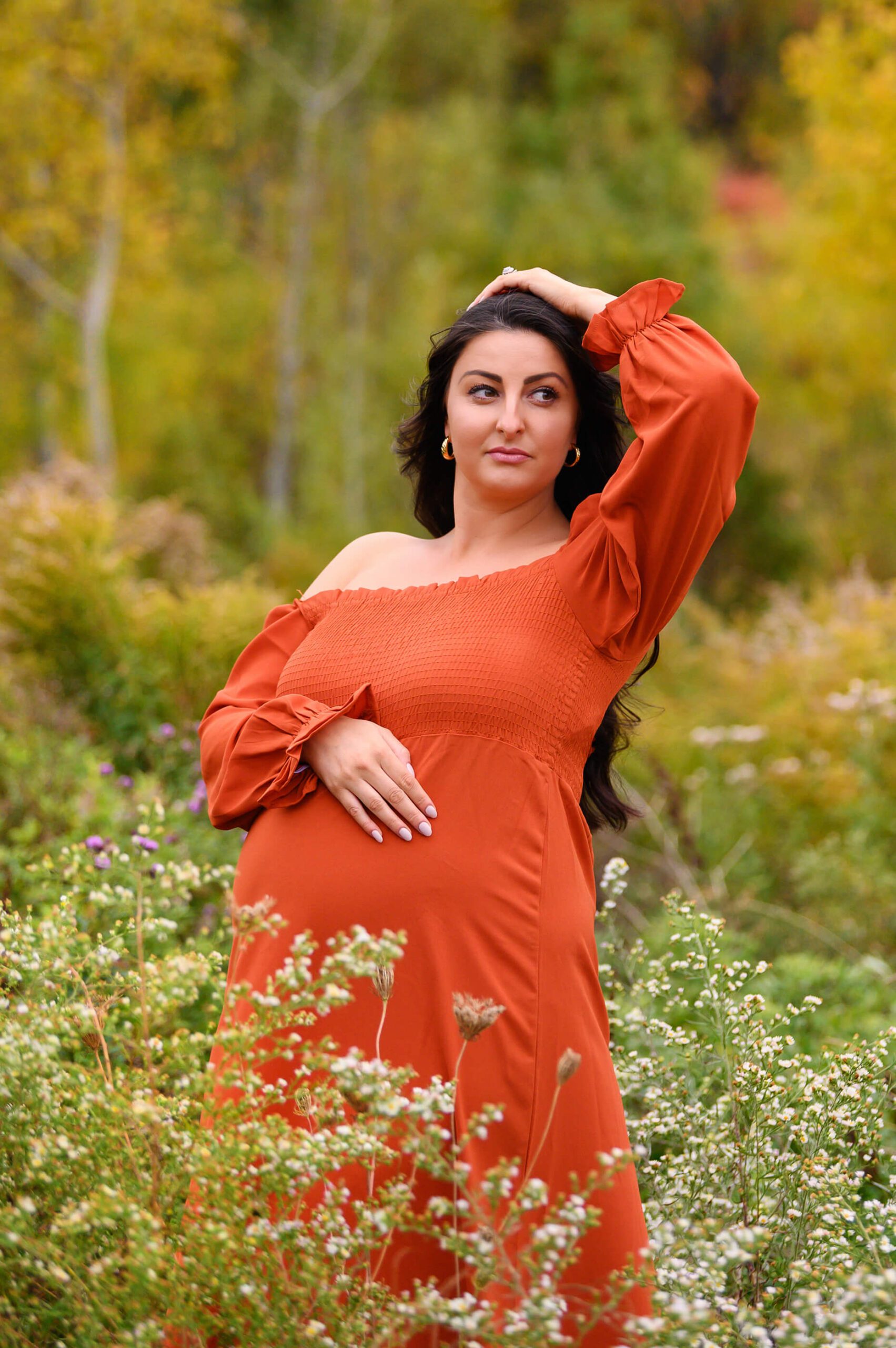 Toronto Maternity Photography Session. Mom is wearing an Orange dress wither her hand on her head looking off into the distance with fall foilage.