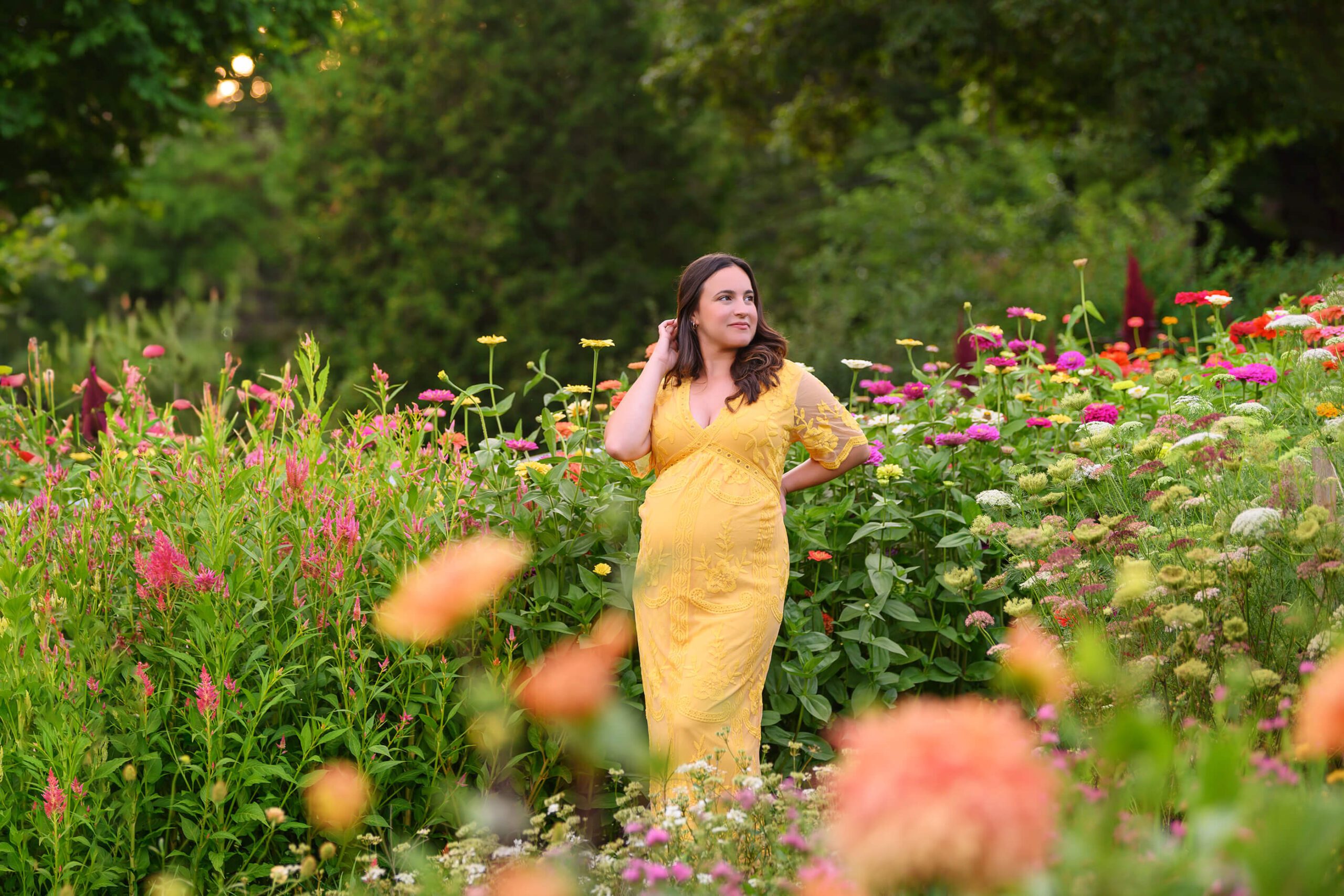mom to be with her hand in her hair in a yellow dress amongst the flowers