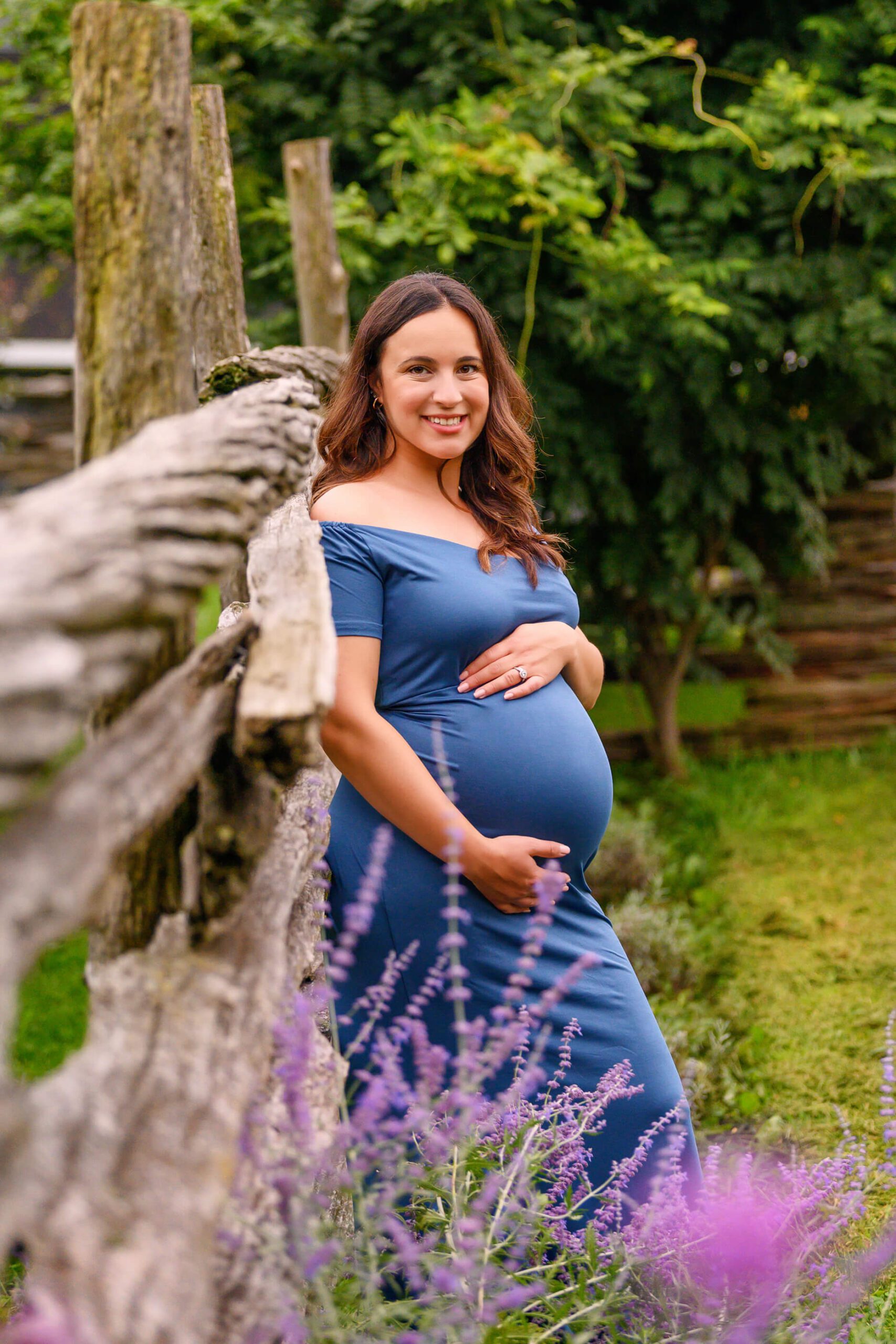 mom leaning against a wood fence holding her belly wearing a blue maternity dress