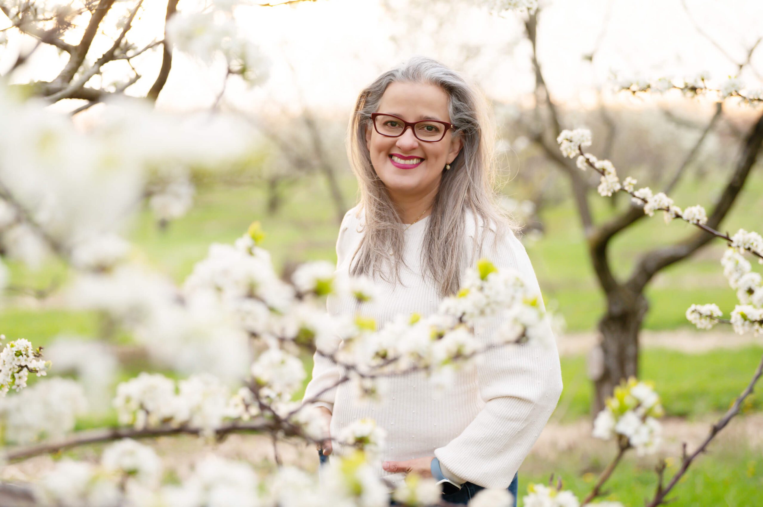 Woman smiling in a white sweater with the sun setting behind in the white cherry blossoms.