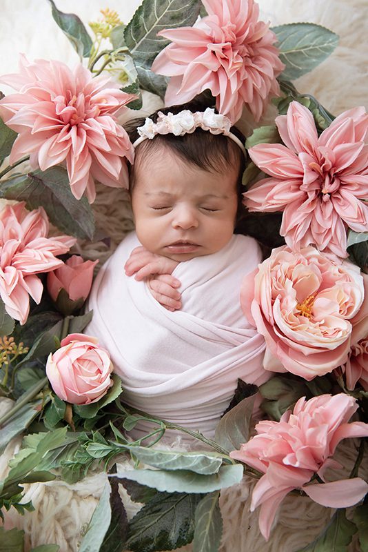 Newborn baby girl lying down wrapped in pink with pink flowers all around her.