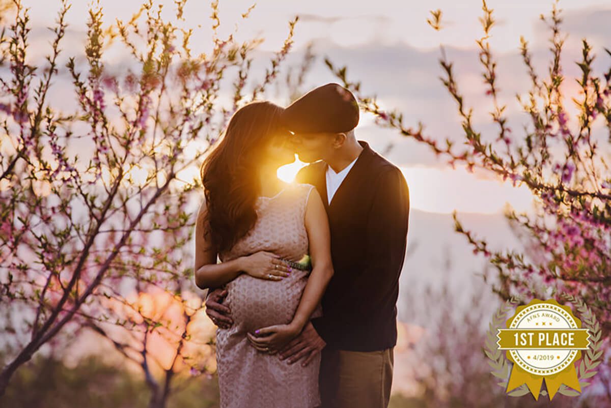 husband and wife kissing with the sun setting behind them for their award-winning maternity cherry blossom photography session.